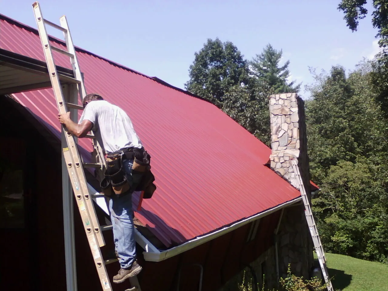 Roofer on a ladder beside a red metal roof and stone chimney — Soffit & Fascia Repair by Roofing Brothers in Saint Ignatius